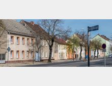 Straße mit einer Reihe von zweistöckigen Wohnhäusern, deren Fassaden in verschiedenen hellen Farbtönen gestrichen sind, wie Beige und Grün. Bäume ohne Laub säumen den Gehweg, daneben Verkehrszeichen und ein Straßenschild mit der Aufschrift „Rudolf-Breitscheid-Straße“.