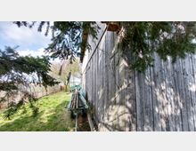 Holzschuppen mit verwitterter Holzfassade, daneben ein grüner Anhänger mit gestapelten Ästen. Im Vordergrund sind Nadelbaumzweige sichtbar, im Hintergrund ein Zaun und Bäume auf einem grasbewachsenen Feld.