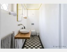 Schmales Badezimmer mit weißen Wänden, schwarz-weiß kariertem Boden, eingelassenem Waschbecken auf einer Holzplatte, wandmontiertem Wasserhahn, großem Spiegel und Toilette im Hintergrund.