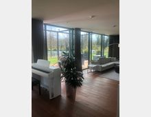 Modernes Wohnzimmer mit großen Glasfronten, die den Blick auf einen Garten mit Bäumen und einen Fluss freigeben. Der Raum ist mit Holzboden, einer grauen Eckcouch, einem weißen Klavier und einem großen Zimmerpflanzentopf ausgestattet.