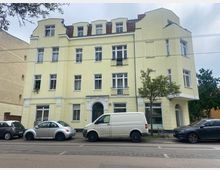 Gelbes, dreistöckiges Wohngebäude mit Mansarddach und mehreren Fenstern, darunter Balkone an der oberen Etage. Vor dem Gebäude befinden sich parkende Autos und ein kleiner Baum, daneben verläuft eine Straßenbahnlinie.