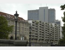 Straßenansicht mit gemischter Architektur: links ein historisches Gebäude mit rotem Ziegeldach und rechteckigen Fenstern, daneben ein moderner Wohnblock mit Balkonen und Betonfassade, im Hintergrund ein Hochhaus mit Glasfassade. Im Vordergrund stehen Laternen und ein Brückengeländer, umgeben von Bäumen.