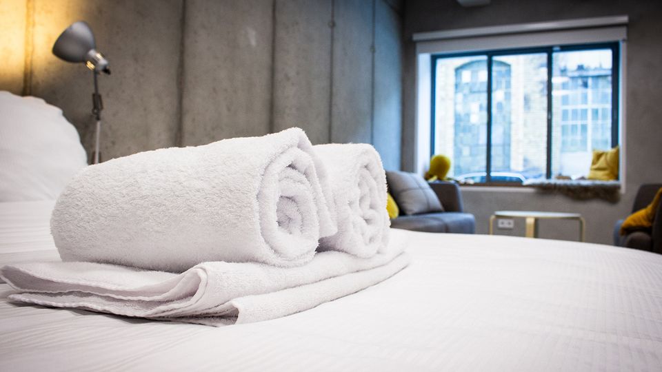 Modernes Hotelzimmer im Industrial-Stil mit Betonwänden, großem Fenster zur Stadt und einem Bett im Vordergrund, auf dem zwei gerollte weiße Handtücher liegen. Im Hintergrund sind ein Sofa mit Kissen, eine Stehlampe und ein kleiner Tisch zu sehen.