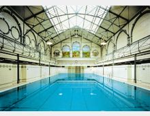 Historisches Schwimmbad mit einem rechteckigen Becken in einem hoheren Saal mit gewölbter Stahl- und Glasdecke, weißen Fliesenwänden mit dekorativen Mustern und umlaufender Galerie. Auf der Stirnseite befinden sich große Fenstermotive mit gemalten Landschaftsbildern.