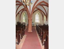 Innenraum einer kleinen Kirche mit roten Backstein-Gewölbebögen und einem zentralen Säulenpfeiler. Auf beiden Seiten befinden sich hölzerne Kirchenbänke, während im Hintergrund zwei hohe, spitzbogige Fenster mit Buntglas und ein einfacher Altartisch zu sehen sind.