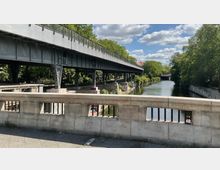 Eine steinerne Brücke mit Metallgeländer führt über einen Fluss, der von Bäumen und Uferbefestigungen mit Graffiti gesäumt wird. Über der Szenerie verläuft eine erhöhte Stahlbahntrasse.