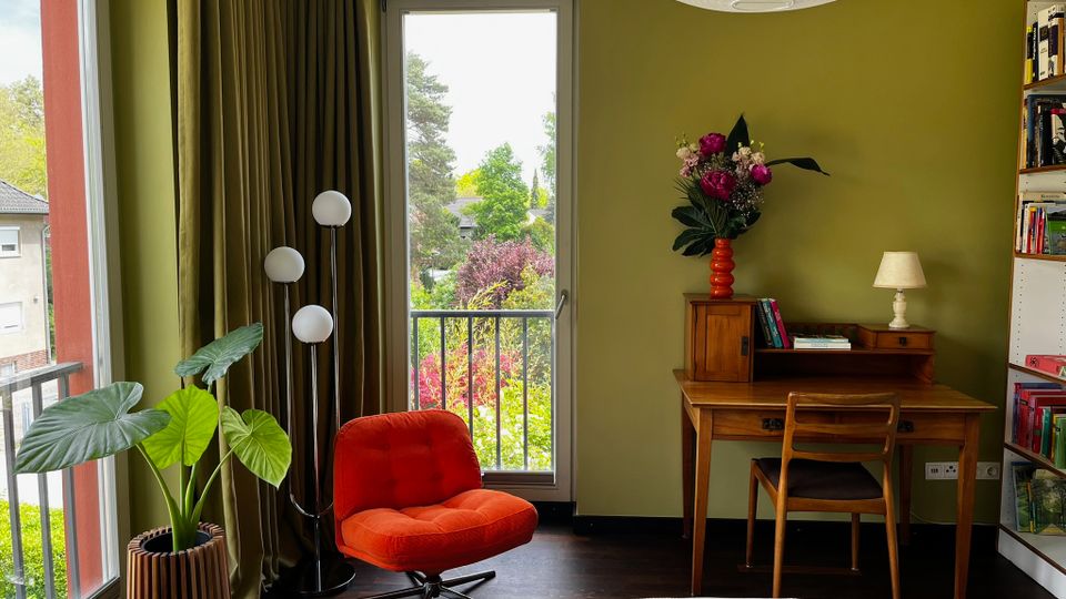 Gemütliches Schlafzimmer mit olivgrünen Wänden, großem Papier-Deckenlampenschirm und dunklem Holzboden; links ein roter Sessel neben einer hohen Stehlampe und einer großen Topfpflanze. Rechts steht ein Holzschreibtisch mit Stuhl, Tischlampe und Blumenvase, daneben ein Bücherregal; durch bodentiefe Fenster und eine Glastür mit Vorhängen blickt man auf einen Balkon und viel Grün im Außenbereich.