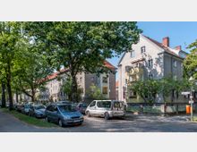 Wohnstraße mit mehrstöckigen Wohnhäusern im Stil der 1920er Jahre, teilweise mit Balkonen und rot gedeckten Dächern. Links sind Bäume, die die Straße säumen, sowie parkende Autos auf einer gepflasterten Fahrbahn sichtbar.