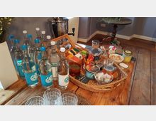 Ein rustikaler Holztisch mit einer Auswahl an Getränken und Snacks: mehrere Wasserflaschen, ein Korb mit Teebeuteln, Gewürzdosen, Zuckerwürfel, Gläser und kleine Schalen. Im Hintergrund ein kleiner runder Holztisch und eine Wasserkanne.