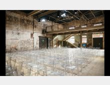 Ein Veranstaltungsraum in einem industriellen Stil mit freigelegten Ziegelwänden, einer Metalltreppe im Hintergrund und einer Galerieebene. Auf dem Boden sind zahlreiche transparente Kunststoffstühle aufgestellt, und die Decke zeigt Stahlträger und Lampen.