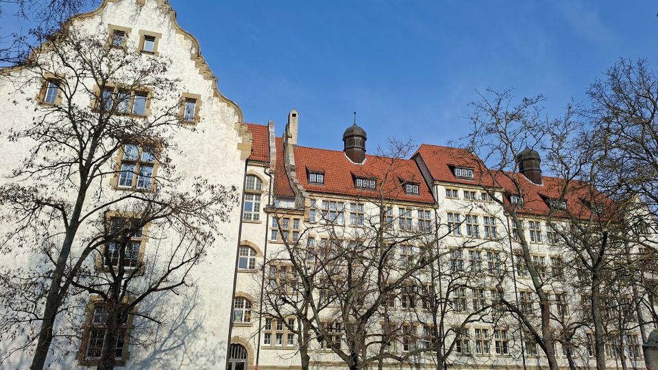 Großes historisches Gebäude mit heller Putzfassade und rotem Ziegeldach, geprägt von geschwungenen Giebeln, vielen Fenstern und kleinen Dachaufbauten mit Türmchen. Im Vordergrund stehen kahle Bäume vor der Fassade unter blauem Himmel.