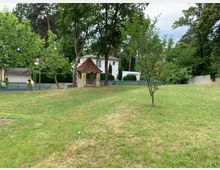 Grünfläche mit leichtem Gefälle, vereinzelt bewachsen mit jungen und älteren Bäumen. Im Hintergrund stehen ein kleines Steinhäuschen mit Ziegeldach und mehrere weiße Wohnhäuser, umgeben von hohen Bäumen und einem Holzzaun.