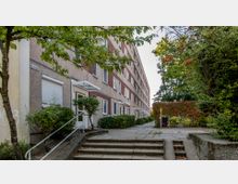 Mehrstöckiges Wohngebäude in einer städtischen Umgebung mit einer Fassade aus Beton und roten Akzenten. Vor dem Gebäude befinden sich ein gepflasterter Gehweg, Treppenstufen, Sträucher und Bäume, die von grünem Laub umgeben sind.