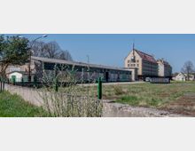 Industrieanlage mit großen, flachen Hallen und einem mehrstöckigen Gebäude im Hintergrund, das ein Satteldach und eine Antenne auf der Spitze hat. Die Umgebung besteht aus Grünflächen, einem Betonzaun und vereinzelten Bäumen.