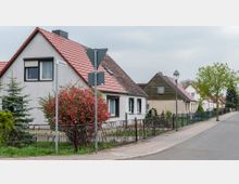 Reihen von Einfamilienhäusern mit Satteldächern in einer ruhigen Wohnstraße. Vor den Häusern befinden sich gepflegte Vorgärten mit Zäunen, einem blühenden Busch und Gehwegbeleuchtung.