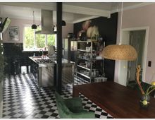 Moderne Küche mit schwarz-weißen Schachbrettfliesen, einer freistehenden Edelstahl-Kochinsel und schwarzen gefliesten Wänden. Im Vordergrund ein Holztisch mit grünem Stuhl, auffälliger Korblampe und Dekoration, im Hintergrund offene Regale und ein großes Fenster mit Blick auf grünes Laub.