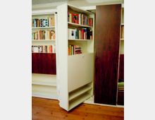 Innenraum mit eingebauten Bücherregalen aus weißem Holz und dunkel furnierten Schranktüren; ein schwenkbares Regal ist geöffnet und zeigt mehrere Reihen mit Büchern. Holzboden und eine niedrige Decke rahmen die Regalanlage ein.