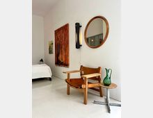 Minimalist bedroom corner with white walls and floor, featuring a low bed, a wooden lounge chair, and a small round metal side table with a green glass vase. Wall decor includes a large orange-toned painting, a smaller artwork, a vertical wall sconce, and a round wooden-framed mirror.