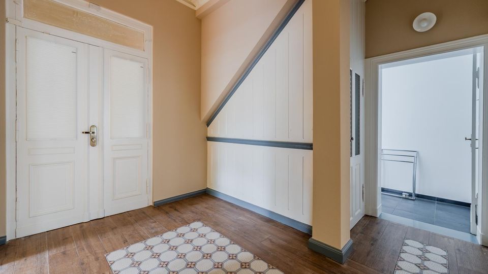 Innenflur eines Altbaus mit hohen Decken, hellen Wänden und dunklem Holzboden mit gemusterten Fliesenläufern. Links eine große weiße Doppeltür, rechts ein Durchgang zu einem hellen Raum; an der Wand ein weißer Treppenunterbau mit dunkler Zierleiste.