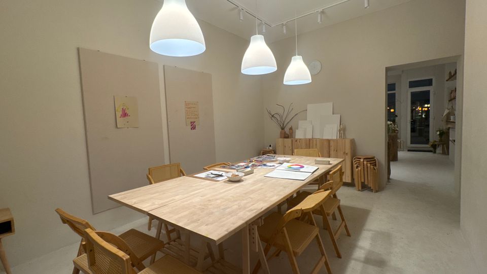 Heller, minimalistischer Workshop- oder Atelierraum mit großem Holztisch und mehreren Holzstühlen, beleuchtet von drei weißen Pendelleuchten. An den hellen Wänden hängen große Akustikpaneele und kleine Bilder; im Hintergrund stehen Holzschränke mit gestapelten Leinwänden und ein Durchgang führt in einen weiteren Raum mit Fenstern.