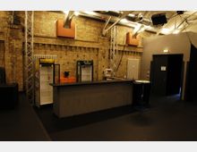 Industrieähnlicher Raum mit einer Theke im Vordergrund, einer freiliegenden Backsteinwand und sichtbaren Metallträgern sowie Lüftungsrohren an der Decke. Im Hintergrund befinden sich zwei Kühlschränke, ein kleiner Tisch und eine Tür.