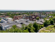 Luftaufnahme eines städtischen Gebiets mit modernen Gebäuden im Vordergrund, darunter ein großer, flacher Bürokomplex mit Glasdächern, umgeben von Bäumen. Im Hintergrund befinden sich ältere, mehrstöckige Backsteingebäude sowie eine weitläufige Wohnbebauung und grüne Landschaft.