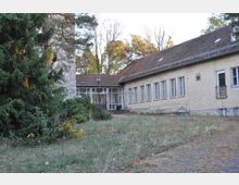 Ein einstöckiges, beige gestrichenes Gebäude mit einem Ziegeldach, großen rechteckigen Fenstern und einer Glastür im zentralen Eingangsbereich; umgeben von Bäumen, Sträuchern und einer Wiese mit vereinzeltem Pflanzenbewuchs.