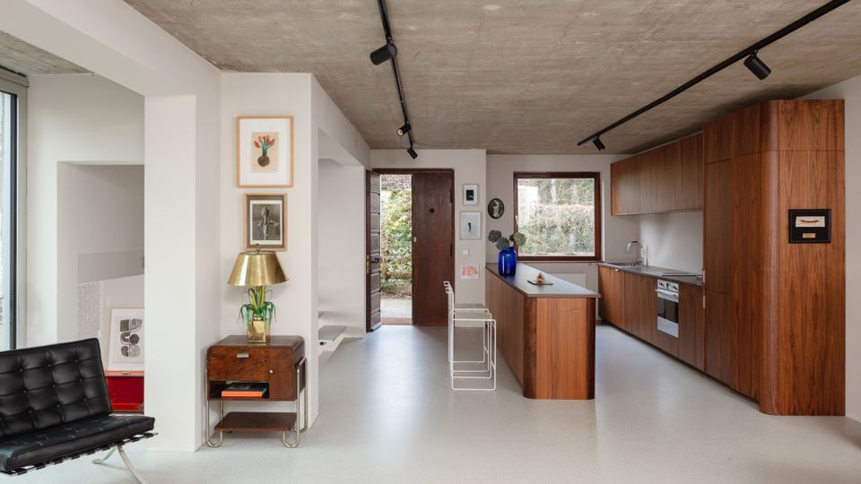 Modern open-plan kitchen and living area with a raw concrete ceiling, white walls, and black track lighting. Warm wood cabinetry and a peninsula with metal stools face a window and open door to greenery, with minimal furnishings and framed artwork along the wall.