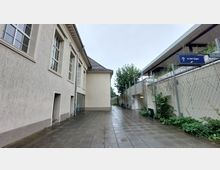 Ein gepflasterter Weg zwischen einem beige verputzten Gebäude mit großen Fenstern auf der linken Seite und einer Betonmauer mit Maschendrahtzaun und Pflanzenbewuchs auf der rechten Seite. Im Hintergrund befindet sich eine Überdachung mit einem Schild, das den Weg "zu den Zügen" weist.