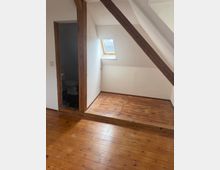 Dachgeschossraum mit schrägen Wänden, sichtbaren Holzbalken und Holzdielenboden; an der Wand sitzt ein kleines Dachfenster. Links führt ein offener Türrahmen in ein kleines Bad mit Toilette und dunklem Fliesenboden.