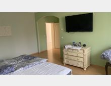 Ein Schlafzimmer mit hellgrünen Wänden, einem Doppelbett vorne links, einer weißen Kommode mit Dekorationsgegenständen darunter ein Fernseher an der Wand und einem offenen Türdurchgang, der in einen anderen Raum führt. Der Boden ist aus hellem Holz.