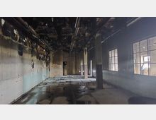 Ein verlassener Raum mit beschädigten Wänden und Decken, teilweise mit verbranntem Material. Die Fensterscheiben an der rechten Seite lassen Licht in den ansonsten dunklen Raum fallen, und der Boden scheint feucht oder nass zu sein.