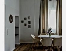 Esszimmer mit einem ovalen weißen Tisch und sechs beigefarbenen Stühlen auf Holzboden. Im Hintergrund befinden sich beige Vorhänge vor einem Fenster, eine Vase mit Blumen auf dem Tisch und an der Wand dekorative Kunstwerke sowie ein runder Wandspiegel.