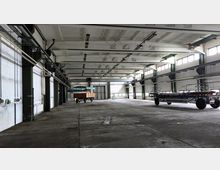 Große, leere Industriehalle mit einer hohen Decke, sichtbaren Stahlträgern und mehreren schmalen Fenstern entlang der Seitenwände. Im Raum befinden sich ein Anhänger mit Holzaufbau und ein weiterer flacher Anhänger.