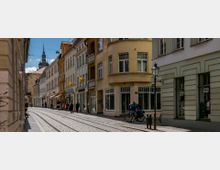 Straßenszene in einer Altstadt mit hellen, mehrstöckigen Gebäuden, die Geschäfte im Erdgeschoss beherbergen. Straßenbahnschienen verlaufen durch die gepflasterte Straße, im Hintergrund ist der Turm einer Kirche mit einer Uhr sichtbar.