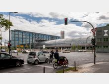 Straßenszene vor dem Berliner Hauptbahnhof, einem modernen Bahnhofsgebäude mit einer großen Glaskuppel und angrenzendem Bürokomplex. Im Vordergrund eine Kreuzung mit Autos, Fahrrädern und Fußgängern, Ampeln und Straßenschildern.