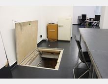 Ein Innenraum mit schwarzem Boden, einer offenen Bodenklappe, die zu einem unterirdischen Bereich führt, und einem alten gelben Schrank sowie einem weißen Kühlschrank an der Wand. Im Hintergrund sind Bürotische und Stühle zu sehen.