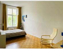 Ein schlicht eingerichtetes Schlafzimmer mit Holzboden, einem großen Fenster mit gelben Vorhängen und einer offenen Aussicht auf grüne Bäume. Im Raum befinden sich ein Bett, ein heller Sessel, ein Nachttisch mit einer Pflanze und moderne Wanddekoration.