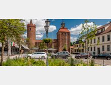 Historischer Marktplatz mit Blumenbeeten im Vordergrund, flankiert von Gebäuden mit roten Ziegeldächern und einer mittelalterlichen Backsteinburg mit zwei Türmen im Hintergrund. Autos parken entlang der Straße, und eine Laterne mit Blumenschmuck steht zentral.