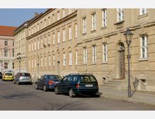 Straße mit einer Reihe von historischen, beigefarbenen Gebäuden mit weißen Sprossenfenstern und Steinverzierungen. Mehrere geparkte Autos sowie Laternenmasten säumen den Gehweg, während im Hintergrund ein weiteres Gebäude mit rosafarbener Fassade sichtbar ist.