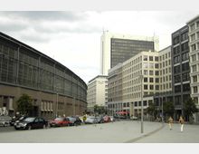 Städtischer Platz mit umliegenden Gebäuden; links ein Backsteingebäude mit großen Fenstern, rechts moderne Bürogebäude. Autos parken entlang der Straße, und einige Personen gehen auf dem offenen, gepflasterten Platz.