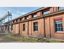 Backsteinfassade eines historischen Industriegebäudes mit großen Fenstern, einem grauen Metallgerüst am linken Bildrand und einer Regenrinne an der Wand. Der Boden im Vordergrund ist unbefestigt und teilweise bewachsen.