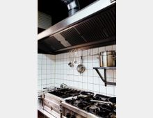 Professionelle Restaurantküche mit Edelstahlherd, mehreren Gaskochfeldern und einer großen Dunstabzugshaube. Weiße geflieste Wände, Kochutensilien an einer Hakenleiste und ein Topf auf einem Regal ergänzen den Arbeitsbereich.