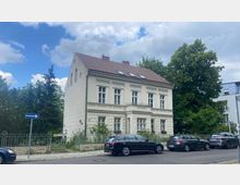 Zweistöckiges weißes Wohnhaus im klassischen Stil mit Satteldach und symmetrischen Fenstern, umgeben von einem Zaun und Gärten. Im Vordergrund sind geparkte Autos und ein Straßenschild für eine Einbahnstraße sichtbar, während sich links und rechts Bäume befinden.