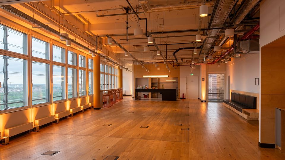 Spacious modern loft-style office or event space with a wide wooden floor, exposed ceiling pipes and suspended lights, and floor-to-ceiling windows along one wall overlooking a cityscape. A small kitchenette and high-top seating area sit at the back, with built-in bench seating along the opposite wall.