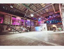 Innenraum einer modernen, umgebauten Industriehalle mit hohen Decken, freiliegender Metallkonstruktion und bunter Beleuchtung. Der Raum zeigt eine Mischung aus Glasfronten, Holzstrukturen, einer Treppe, einer großen Rutsche und offenen Arbeits- oder Veranstaltungsbereichen.