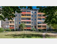 Wohnblock mit vier Stockwerken, grauer Fassade und Balkonen in Gelb-, Orange- und Rottönen, umgeben von einem begrünten Hof mit Wäscheleinen, Rasen und vereinzelten Bäumen. Ein schmaler Weg führt durch den Hof, der von Sträuchern und Bäumen eingerahmt wird.