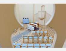 Blick in einen hellen Raum mit kirchlicher Atmosphäre, eingerichtet für eine Zeremonie. Zu sehen sind eine Holzbank mit weißen Überzügen und Blumen, umgeben von mehreren Stuhlreihen mit blauen Polstern, auf einem Boden aus hellen Fliesen und Teppichen.