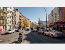 Straßenszene in einer städtischen Umgebung mit mehrstöckigen Wohngebäuden. Links sind helle Altbauten mit Balkonen zu sehen, während rechts ein buntes Gebäude mit Gastronomie im Erdgeschoss steht; parkende Autos und vereinzelte Bäume säumen die Straße.