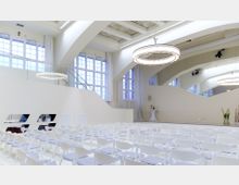 Moderner Veranstaltungsraum mit weißen Stühlen in Reihen, großen Fenstern mit viel Tageslicht und geschwungenen Architekturdetails. Die helle Raumgestaltung wird durch eine dekorative Hängelampe und Spiegelwände ergänzt, die den Raum optisch vergrößern.